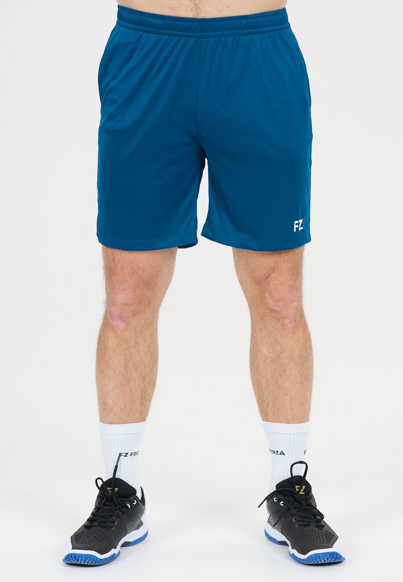 Manliga ben iklädda blå sportshorts, vita crew-strumpor och svarta sportskor som står på vit bakgrund.
