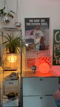 Estante de metal com plantas, uma lâmpada redonda laranja, bola de discoteca e várias obras de arte. Armário branco com um detalhe em madeira horizontal na parte superior.