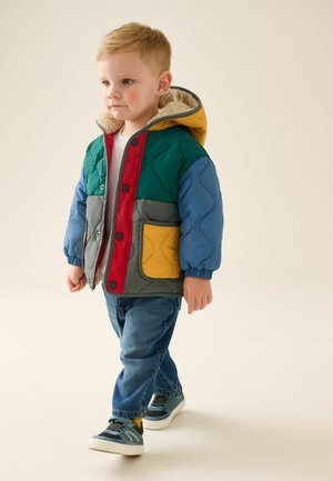 Young child walking wearing a colorful quilted jacket with a hood, blue jeans, and sneakers on a light background.