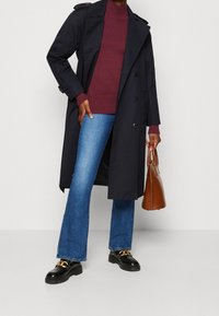 Manteau trench navy à double boutonnage, pull en tricot bordeaux, jeans bleu évasés, mocassins noirs avec des accents dorés et sac à main marron.