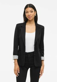 Frau mit langen dunklen Haaren, die silberne Creolen, ein schwarzes Blazer mit aufgerollten gestreiften Ärmeln, ein weißes Oberteil und schwarze Hosen trägt, vor einem weißen Hintergrund.