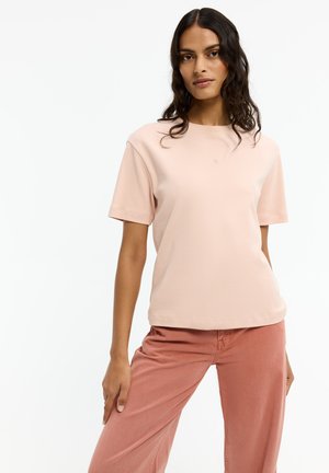 Frau mit langem dunklem Haar, die ein hellrosa kurzärmeliges Hemd und gedämpfte rosafarbene Hose trägt, vor einem einfachen weißen Hintergrund steht.