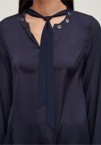 Navy blouse with a front neckline tie, grommet accents, and long sleeves. Made of smooth, lightweight fabric with a soft sheen.