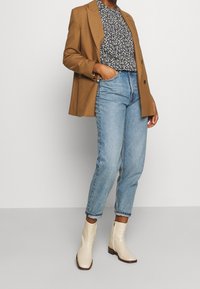 Blazer marron sur un blouse à fleurs, associé à un jean taille haute bleu et des bottines beiges brillantes avec des zips sur le côté.