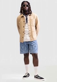 Chaqueta de botones marrón claro, camiseta blanca, pantalones cortos con patrón de llamas azules, calcetines blancos, zapatillas negras, gafas de sol redondas y una bufanda azul.