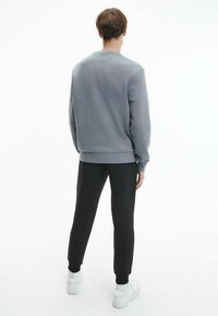 Felpa grigia con scollatura rotonda, abbinata a pantaloni neri aderenti e sneakers bianche, vista da dietro su uno sfondo bianco.