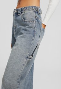 Jeans de mezclilla azul claro con una cintura alta, presillas para cinturón, bolsillo lateral y un corte relajado. La tela tiene un acabado texturizado con costuras visibles.