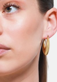 Orecchino a cerchio in oro con un design elegante e curvo. Superficie lucida e riflettente, caratterizzata da una texture liscia e una forma affusolata.