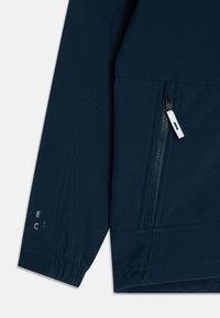 Veste softshell bleu marine, avec un coin, featuring une poche zippée avec un tirette blanche, des poignets élastiques et un détail de logo en relief sur la manche.