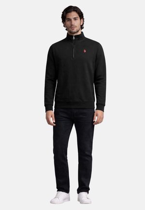 Man standing wearing a black quarter-zip pullover with red logo, black jeans, and white sneakers against a plain white background.