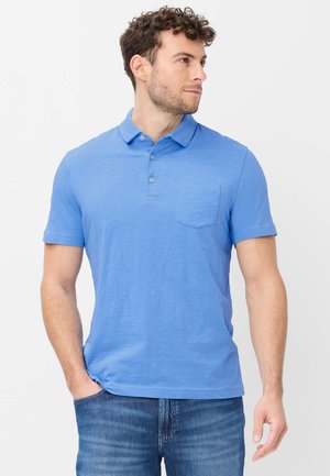 Man met krullend haar, gekleed in een lichtblauw poloshirt en blauwe spijkerbroek, staand met één hand in de zak, kijkt naar rechts.