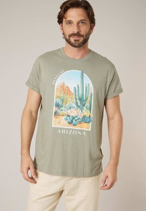 Homme portant un t-shirt vert clair aux manches retroussées avec une scène du désert de l'Arizona, incluant des cactus et des formations rocheuses, assorti à un pantalon beige.