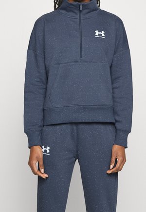 Osoba nosząca granatową, nakrapianą bluzę Under Armour z suwakiem do ćwiartki i pasujące spodnie dresowe, z widocznym logo na piersi i udzie.