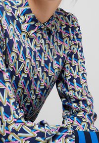 Kauluksellinen paita, joka on valmistettu sileästä kankaasta ja jossa on toistuva "LOVE"-kuvio sinisenä, vaaleanpunaisena ja kultaisena laivastonsinisellä pohjalla. Näkyvissä olevat napit ja kalvosimet.