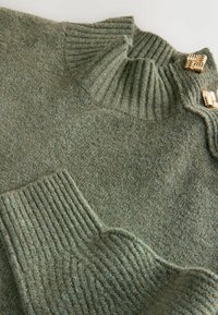 Strickpullover aus weichem grünem Stoff mit geripptem Kragen und Manschetten; verfügt über goldene Hardware-Details am Halsausschnitt für zusätzlichen Akzent.