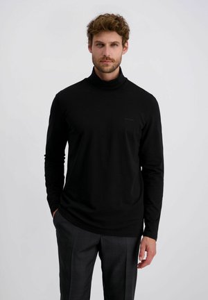 Mann mit lockigem Haar, der einen schwarzen Rollkragenpullover und dunkelgraue Hose trägt, steht vor einem schlichten hellen Hintergrund.