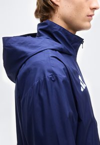 Πλαϊνή όψη ενός ατόμου που φοράει μια μπλε σκούρα κουκούλα με λευκό λογότυπο στο στήθος, μπροστά σε ένα απλό φόντο.