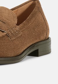 Καφέ καστόρινες μοκασίνια με λεπτομέρεια υποδοχής πεννιού, χαμηλό σωρευμένο τακούνι και υφή στην επιφάνεια, με διαμορφωμένο σχήμα και λείο εσωτερικό επένδυση.