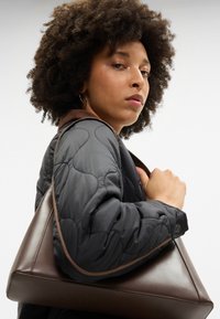 Rjava usnjena torbica v obliki trikotnika z gladko teksturo, kontrastnim šivom in quiltirano črno jakno z teksturirano površino.