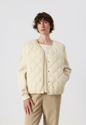 Persona con i capelli corti e ricci che indossa un gilet trapuntato beige sopra un maglione crema e pantaloni marrone chiaro a coste, in piedi davanti a uno sfondo semplice.