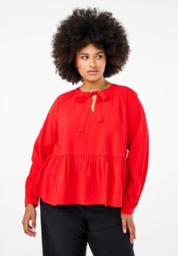 Blusa rossa con maniche lunghe, dalla vestibilità ampia, orlo arricciato e dettaglio a fiocco nel colletto. Realizzata in tessuto leggero.