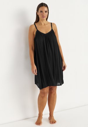 Kobieta stojąca boso, ubrana w luźną czarną sukienkę bez rękawów sięgającą kolan na białym tle.