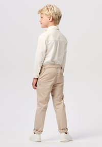 Chemise boutonnée de couleur claire à manches longues, associée à un pantalon beige fuselé et des baskets blanches, mettant en avant un design simple et une coupe décontractée.