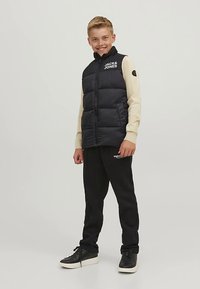 Črna puhasta telovnik z logotipom na prsih, bež majica z dolgimi rokavi, črne trenirke in črni športni copati na stoječem otroku.