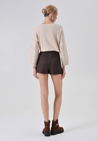 Donna con capelli biondi raccolti in uno chignon, che indossa un maglione beige con maniche paillettate, pantaloni corti marrone scuro e stivaletti alla caviglia marroni, in piedi su uno sfondo bianco.