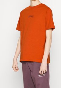 Oranje T-shirt met korte mouwen van katoen, voorzien van een ronde hals en een zwart "new balance" logo op de borst. Casual, losse pasvorm.