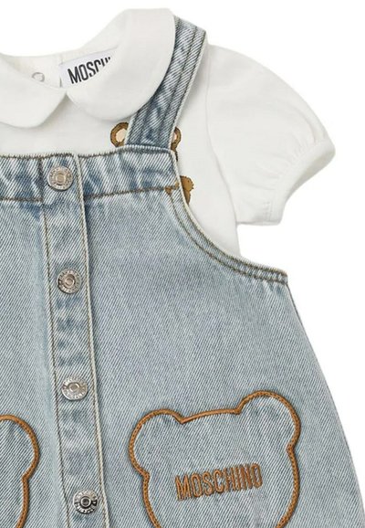 Abito in denim a salopette di un colore azzurro chiaro, con bottoni argentati e contorni di orsetti di peluche marroni sulle tasche. Camicia bianca con colletto rotondo indossata sotto.