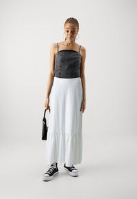 Fekete farmer crop top vékony pántokkal, fehér csipkés maxi szoknya és fekete sneaker. A modell egy kis fekete táskát tart.