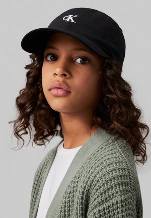 Nooruk lokkis juustega, musta mütsi ja "cK" logoga, helerohelist kudumit kardiganit ja valget särki kandmas, halli taustaga.