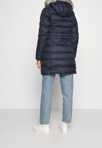 Abrigo acolchado azul marino con capucha forrada de piel, mangas largas y textura acolchada. Se lleva sobre jeans azul claro y zapatillas blancas.