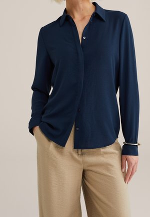 Vrouw die een marineblauw overhemd met lange mouwen draagt, aan één kant in beige gestructureerde broek gestopt, met één hand in de zak en een armband om de pols.