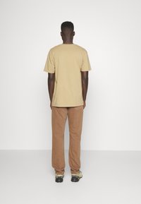 Béžové bavlněné tričko, tanové manšestrové kalhoty a černé sportovní boty. Pohled zezadu, ukazující jednoduchou siluetu a barvy outfitu.