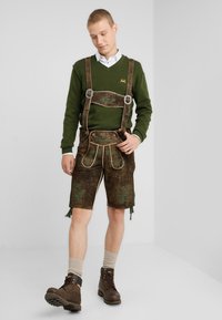 Braune Lederhosen mit grüner floraler Stickerei, braunen Hosenträgern und einem grünen Pullover, der über einem weißen Hemd mit Kragen getragen wird. Braune Stiefel.