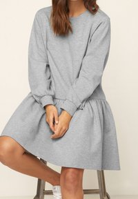 Femme assise sur un tabouret portant une robe sweatshirt à manches longues gris clair et des baskets blanches, les mains posées sur ses genoux.