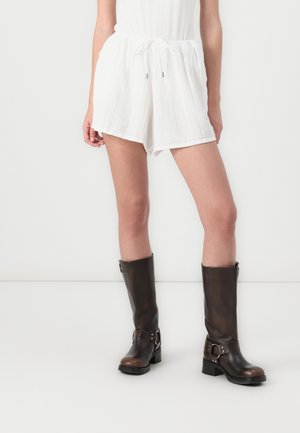 Shorts blancs à cordon de serrage en tissu texturé, assortis à des bottes en cuir marron montantes avec des accents de boucle et une semelle plate.