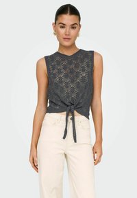 Un top sans manches en tricot gris avec un motif en dentelle, présentant un détail noué à l'avant. Associé à des pantalons taille haute de couleur claire.