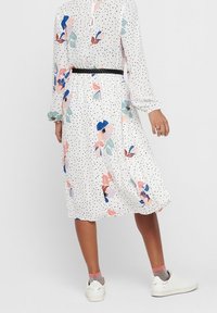 Nainen, jolla on päällä valkoinen midi-mekko mustilla pilkuilla ja värikkäillä kukkakuosisilla yksityiskohdilla, yhdistettynä valkoisiin lenkkareihin ja vaaleanpunaisiin sukkiin.