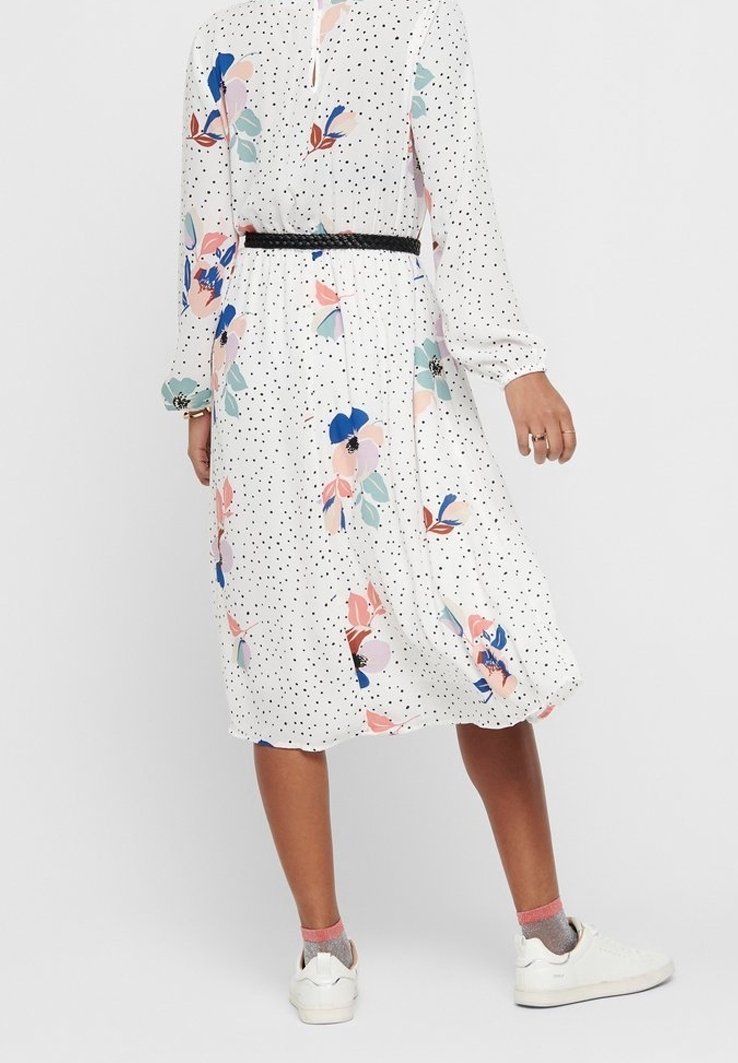 Nainen, jolla on päällä valkoinen midi-mekko mustilla pilkuilla ja värikkäillä kukkakuosisilla yksityiskohdilla, yhdistettynä valkoisiin lenkkareihin ja vaaleanpunaisiin sukkiin.