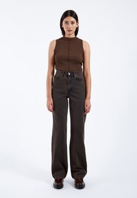 Braun gestreifter ärmelloser Oberteil mit hohem Kragen kombiniert mit hoch taillierten dunkelbraunen, ausgestellten Jeans. Das Model steht zentriert vor einem weißen Hintergrund.