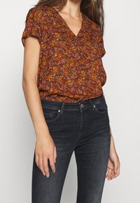 Top de manga corta con escote en V en un patrón floral con tonos naranja, amarillo y azul, combinado con jeans de mezclilla oscuros. Tejido suave y ligero.