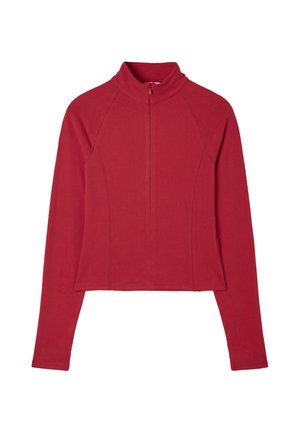 Jeune femme debout portant un haut rouge à manches longues, une ceinture rayée, un pantalon blanc évasé et des ballerines blanches sur un fond gris.