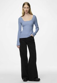 Šviesiai mėlynas raukšlėtas ilgomis rankovėmis viršus su kvadrato apykakle, derinamas su juodomis plačiomis kelnėmis ir juodais batais. Matoma tekstūruota medžiagos detalė.