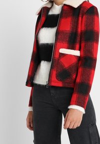 Rød og sort ternet jakke med en cremefarvet imiteret pelskrave og lomme, kombineret med en hvid og sort stribet sweater. Tekstureret stof.