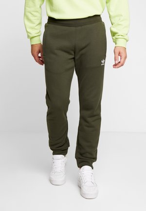 Homme portant un pantalon de survêtement Adidas vert olive, des baskets blanches et un sweat-shirt jaune clair, debout sur un sol et un fond blancs.