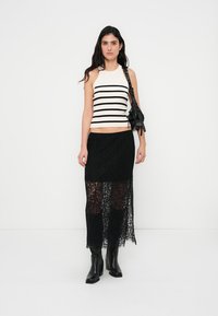 Sort maxi-nederdel i blonder lagdelt over en tætsiddende hvid tanktop med sorte striber. Modellen bærer sorte ankelstøvler og holder en sort taske.