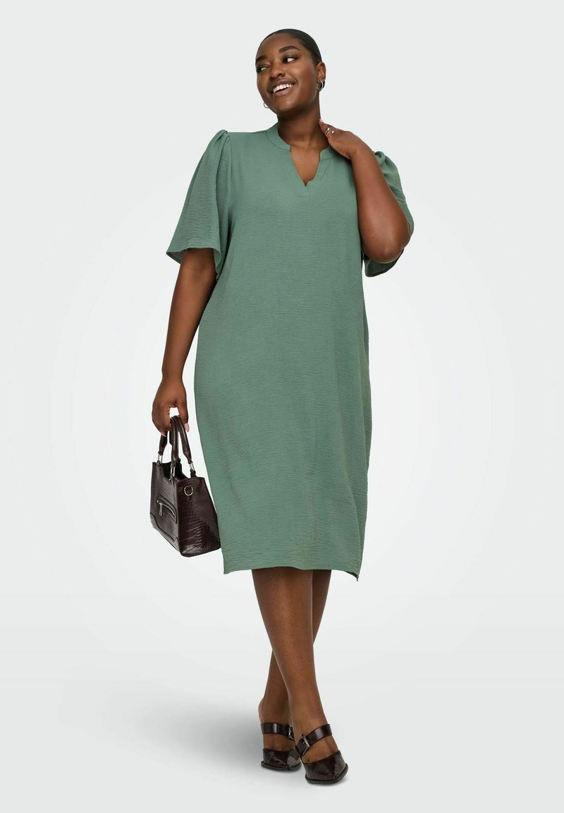 Femeie în rochie verde până la genunchi, cu mâneci scurte bufante, ținând o geantă maro închis, zâmbind și privind în lateral.